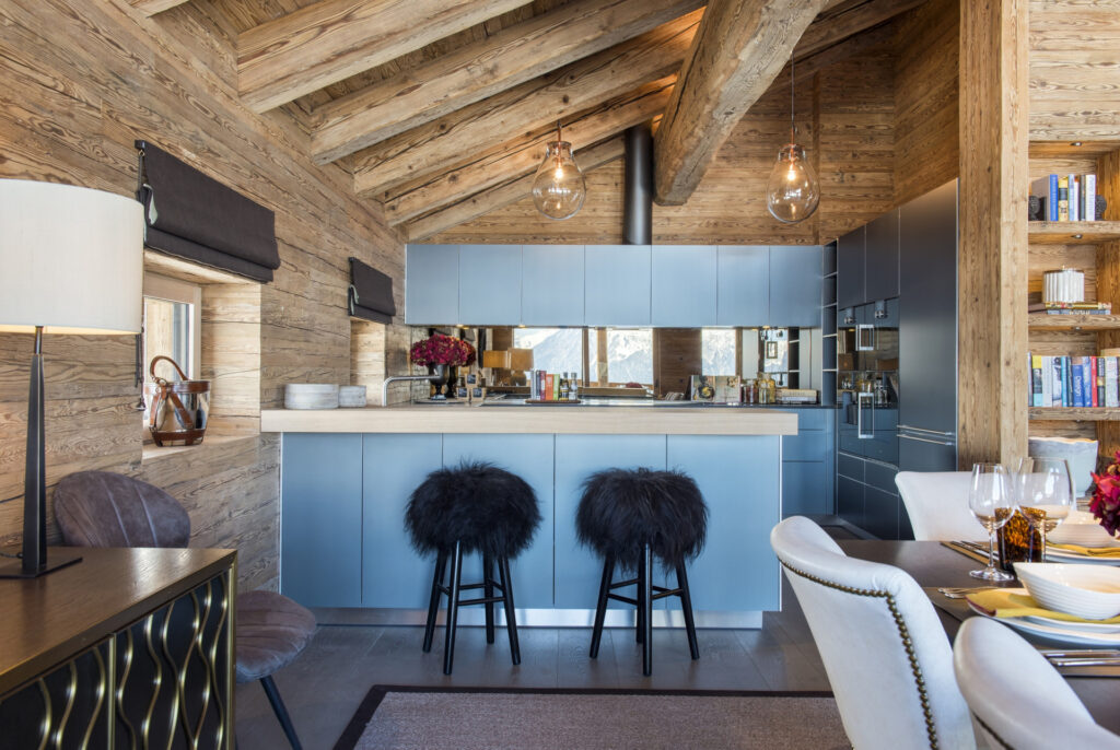 A modern rustic kitchen with light wood walls and ceiling, blue cabinets, a wooden countertop, two black furry bar stools, and glass pendant lights. There is a dining table set for a meal and shelves with books. David Pearson Travel