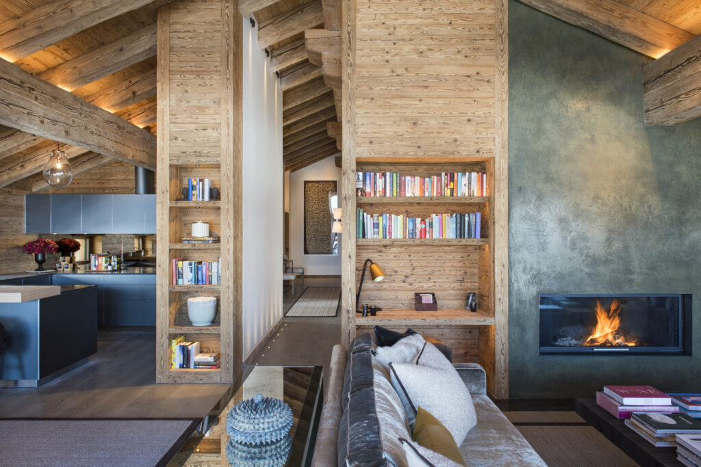 A cozy living room with wooden walls and ceiling, built-in bookshelves, a modern fireplace, and a sofa. The open-plan space connects to a stylish kitchen and features warm lighting and contemporary decor. David Pearson Travel
