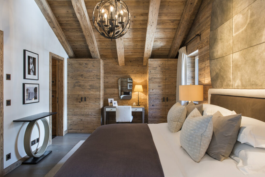 A cozy bedroom with rustic wooden beams, a large bed with gray and beige pillows, a bedside lamp, a modern chandelier, a desk with a chair and lamp, and framed art on the walls. Natural light enters through a window. David Pearson Travel