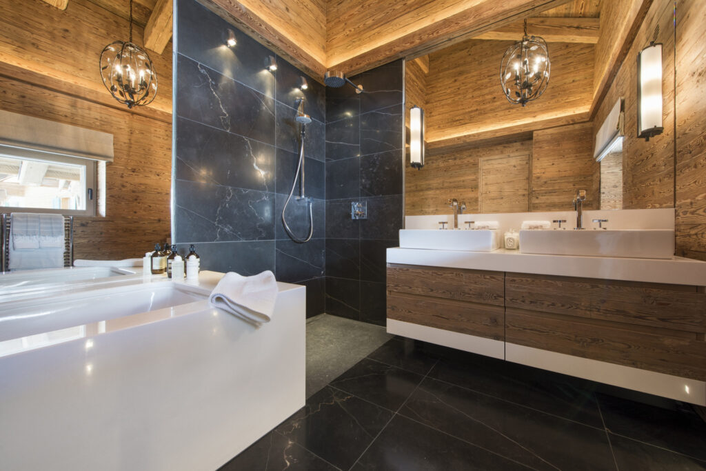 Modern bathroom with wood-paneled walls and ceiling, large black tile shower, double sink vanity with wood cabinets, white bathtub, and elegant lighting fixtures. Natural light enters through a window. David Pearson Travel