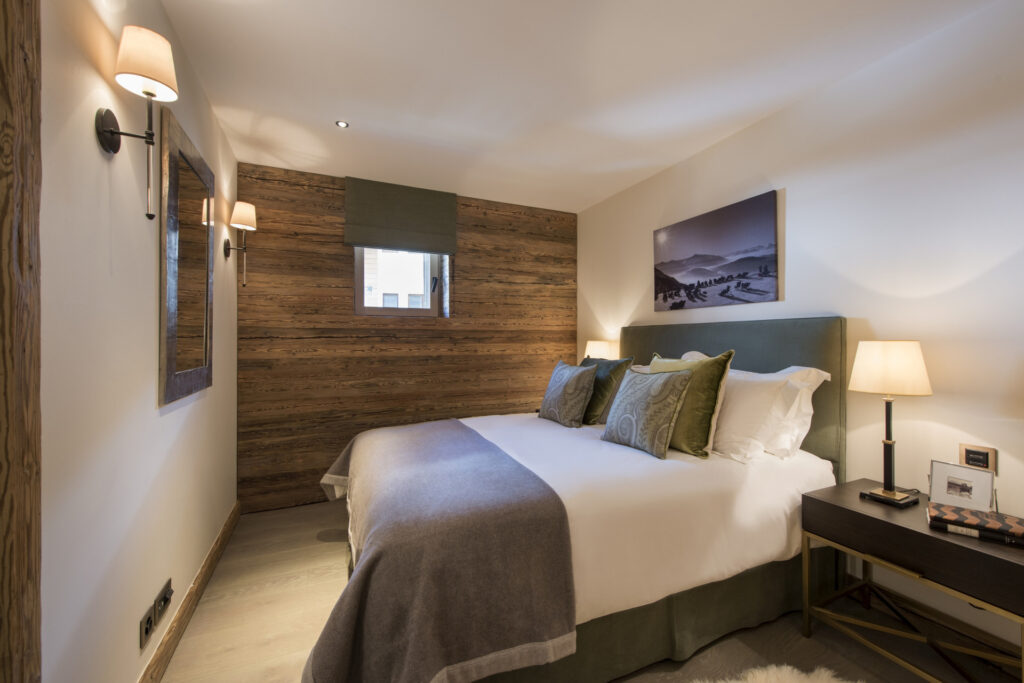 A cozy bedroom with a large bed, green and beige pillows, soft gray blanket, wooden accent wall, bedside tables with lamps, framed photos, a wall mirror, and a small window with a green shade. David Pearson Travel