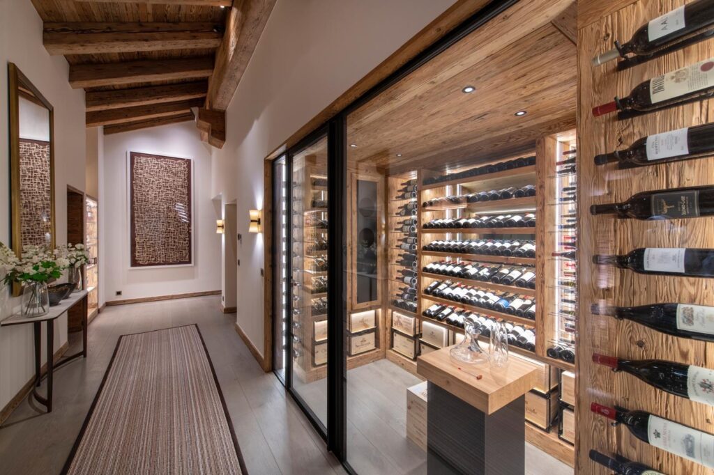 A modern hallway with rustic wooden beams features a glass-enclosed wine cellar filled with bottles, a small wooden table with glasses, and elegant decor including flowers, artwork, and mirrors. David Pearson Travel
