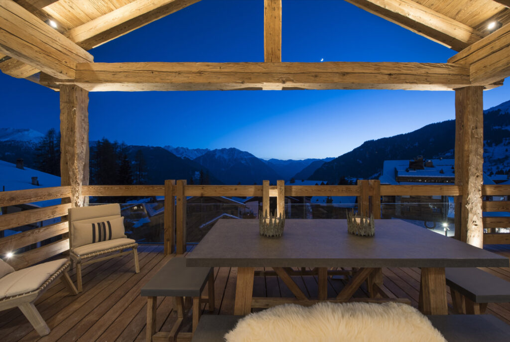 A cozy wooden terrace with a dining table and benches overlooks snow-covered mountains and rooftops at dusk, under a clear, deep blue sky with a crescent moon. David Pearson Travel