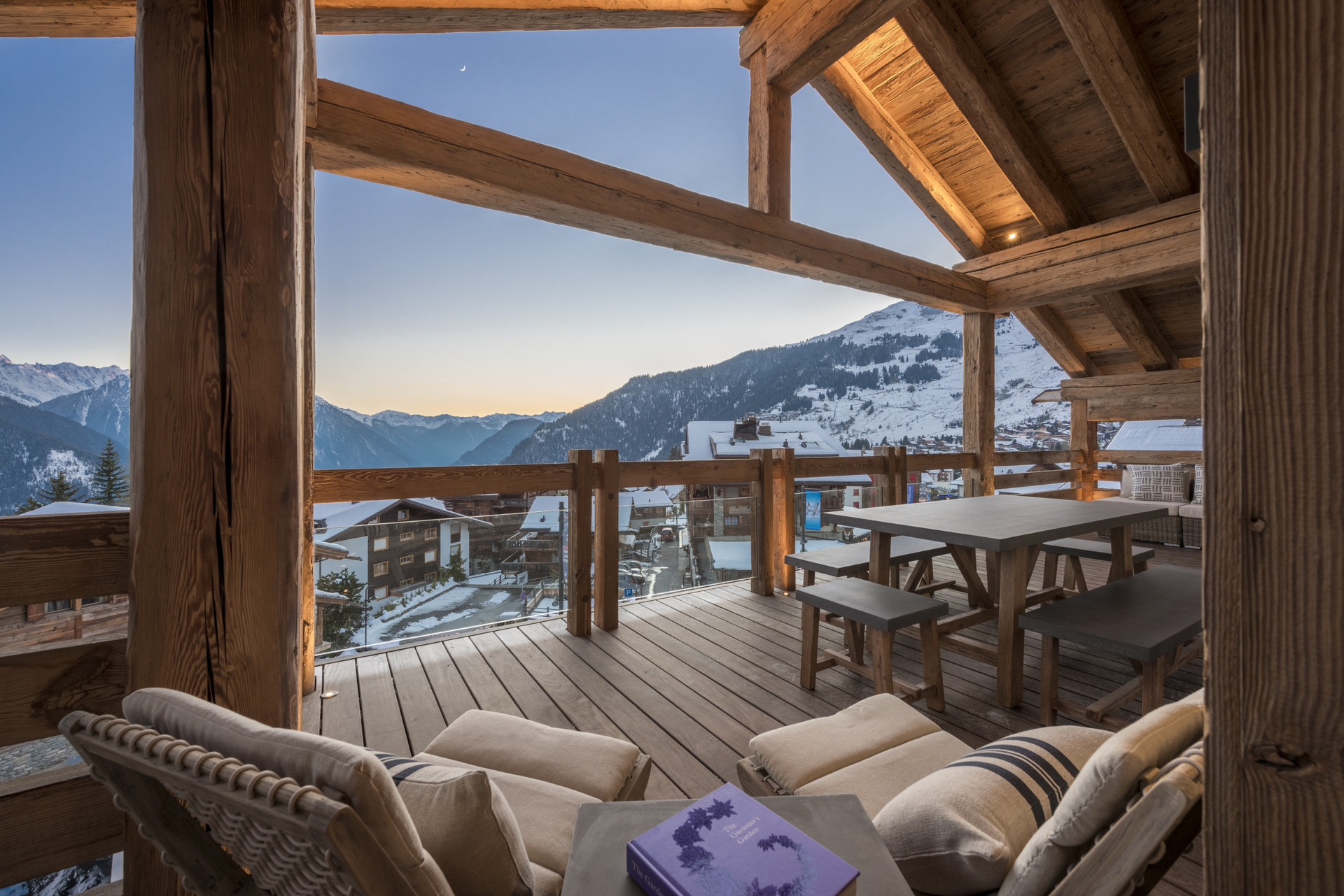A cozy wooden balcony with lounge chairs, a table, and benches overlooks snow-covered mountains and a village at sunset, under a clear sky. David Pearson Travel