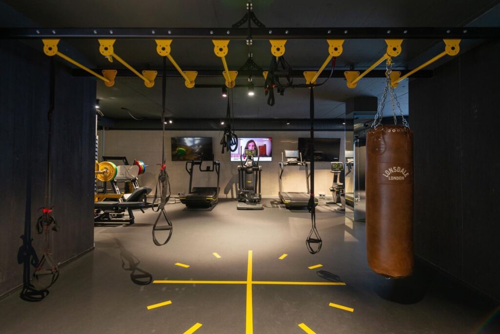 A modern gym with fitness equipment including treadmills, a punching bag, suspension trainers, a weight bench, and a yellow monkey bar structure. A TV screen is visible in the background. The floor has yellow markings. David Pearson Travel