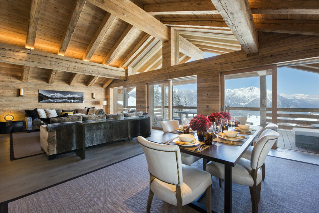 A spacious, luxurious chalet living and dining area with wooden beams, a plush seating area, and a dining table set for six. Large windows reveal a snowy mountain landscape and blue sky outside. David Pearson Travel
