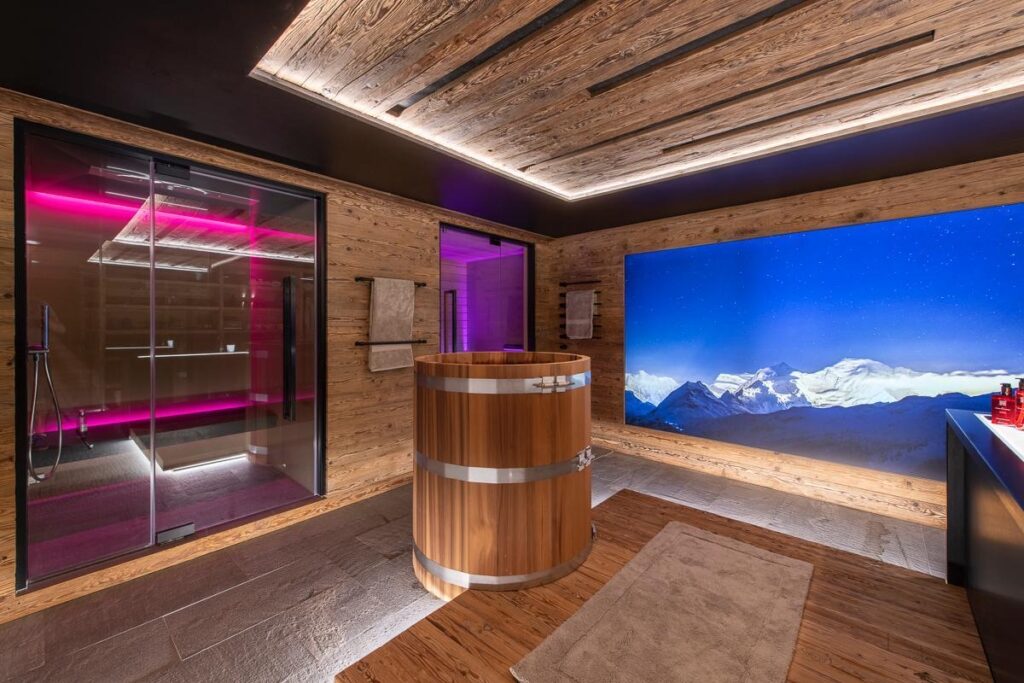 A modern spa room with wood-paneled walls, a round wooden soaking tub, a glass-walled sauna with pink lighting, towels, and a large digital window displaying a scenic mountain view at night. David Pearson Travel