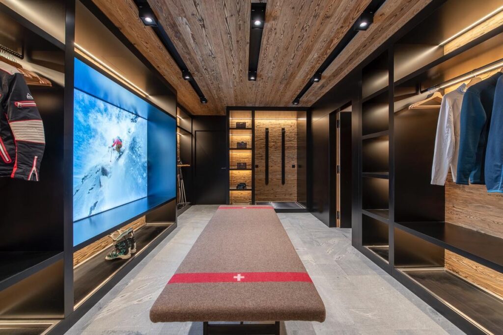 A modern, organized ski locker room with wooden ceiling and shelves, a large bench in the center, hanging ski gear and jackets, cubbies with boots, and a wall-mounted screen displaying a skier on a snowy mountain. David Pearson Travel