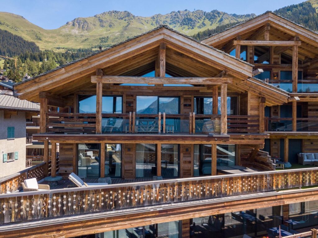 A large wooden chalet with glass balconies and alpine-style architecture stands in front of green hills and mountains under a clear sky. David Pearson Travel