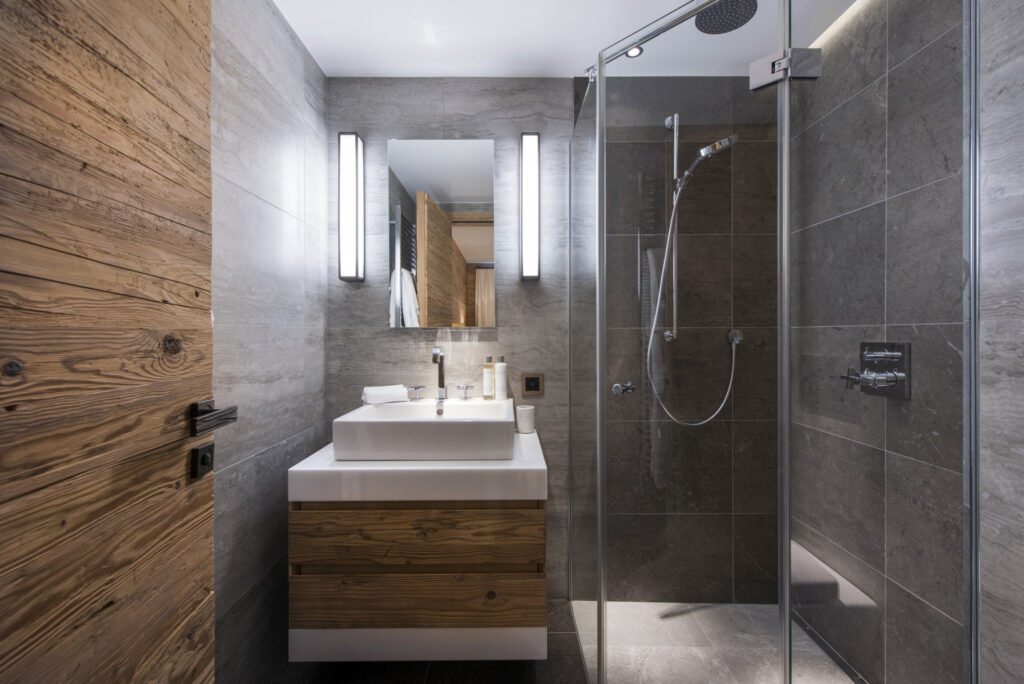 Modern bathroom with gray stone tile walls, a wooden vanity with a white sink, a large mirror with vertical lights on each side, and a glass-enclosed shower with a rainfall showerhead and handheld sprayer. David Pearson Travel