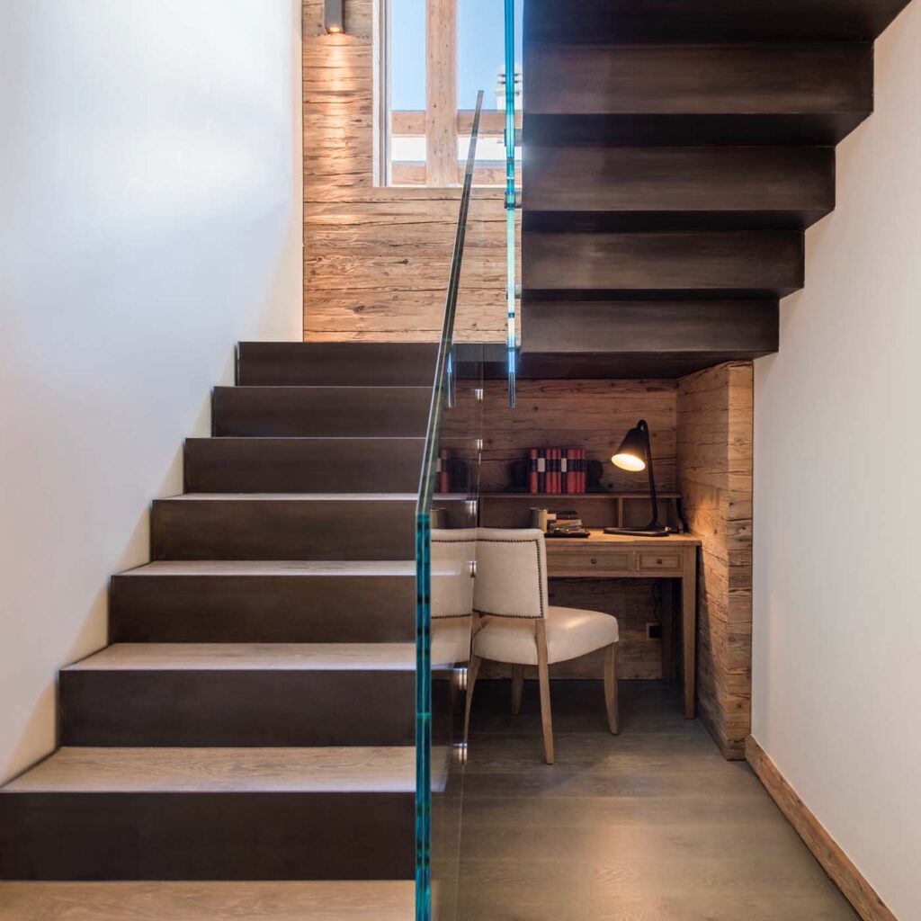 A modern staircase with glass railing leads upward; beneath it is a small workspace featuring a wooden desk, a chair, books, and a lit desk lamp, set against wood-paneled walls. David Pearson Travel