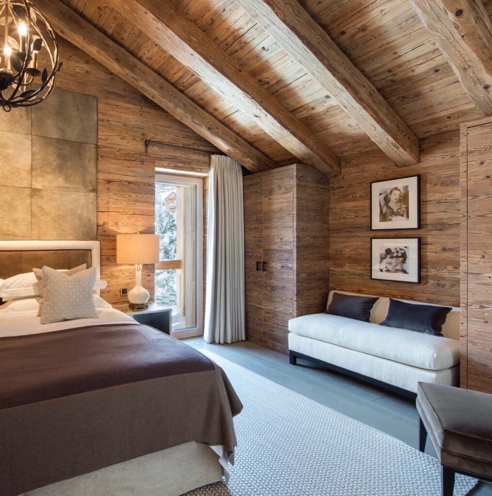 Cozy bedroom with rustic wooden walls and ceiling, a large bed with neutral bedding, a white bench, framed photos on the wall, and natural light coming through a window with curtains. David Pearson Travel