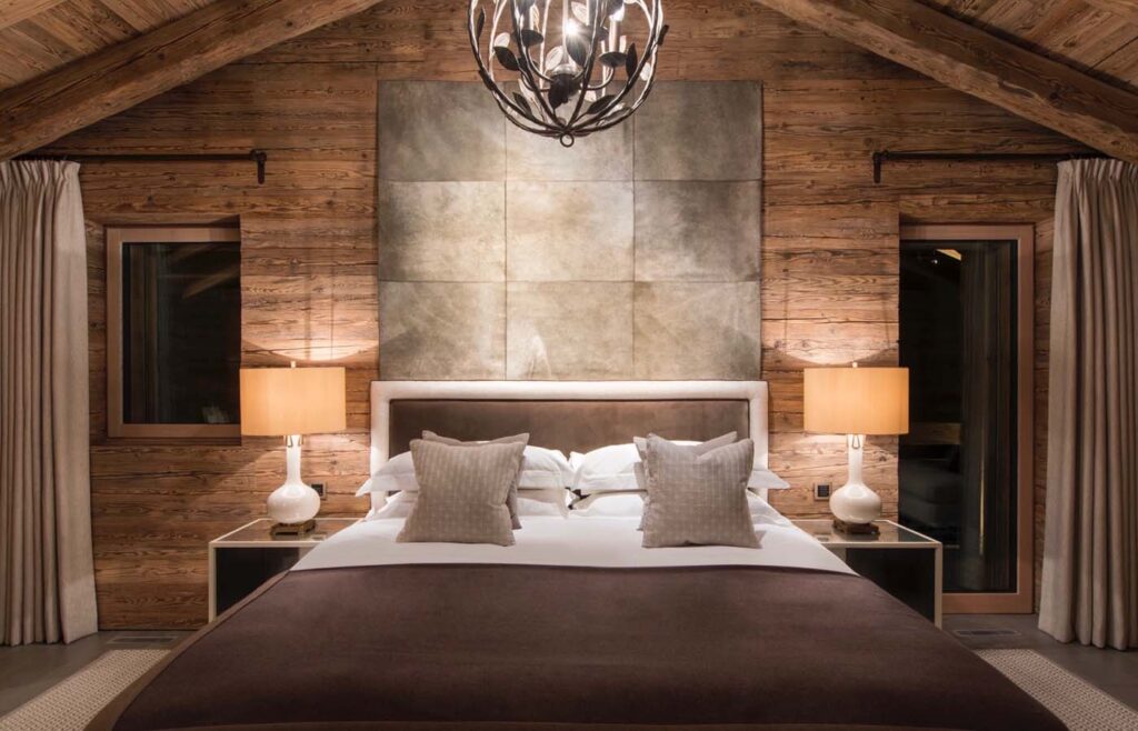 A cozy bedroom with rustic wooden walls, a large bed with white linens and brown throw, two beige pillows, matching bedside tables with lamps, curtains, and modern light fixture hanging from a vaulted ceiling. David Pearson Travel