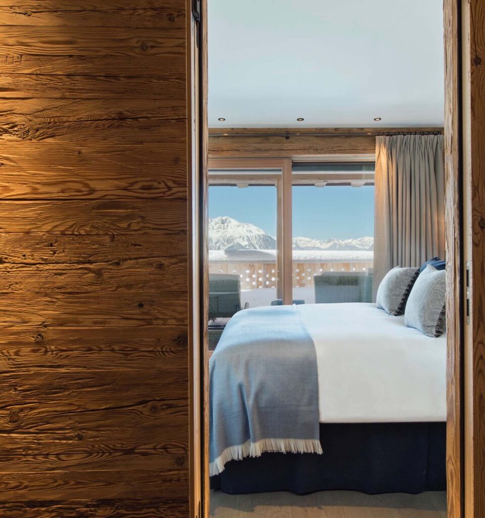 A cozy bedroom with wooden walls, a neatly made bed with blue and white bedding, and a window offering a scenic view of snow-capped mountains under a clear sky. David Pearson Travel