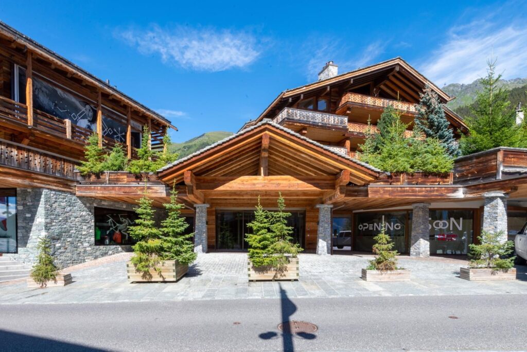 Chalet-style building with stone and wood facade, large overhanging roof, and several potted evergreen trees in front. Mountains and blue sky are visible in the background. David Pearson Travel
