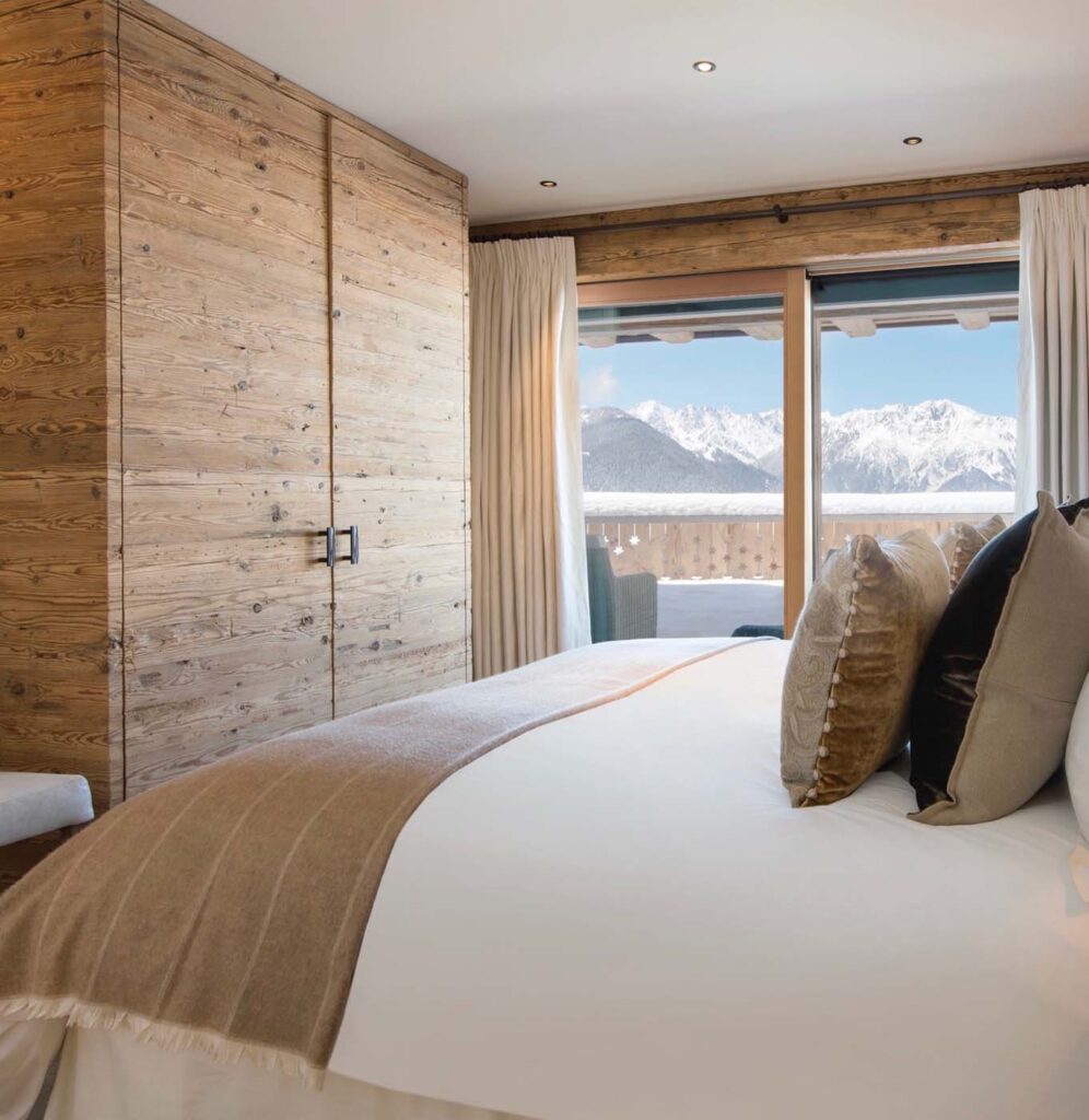 A cozy bedroom with a large bed, wooden wardrobe, and light curtains. A balcony with a scenic view of snowy mountains is visible through the glass doors. David Pearson Travel