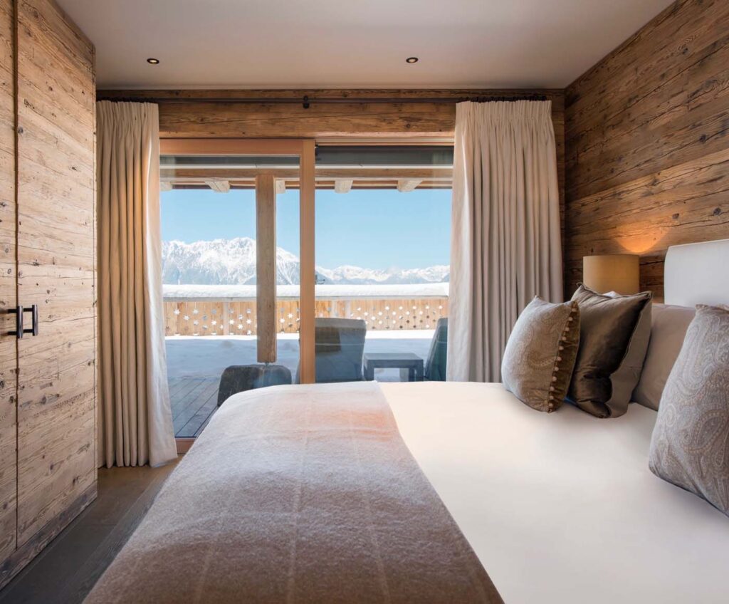 A cozy bedroom with wood-paneled walls, a neatly made bed with neutral-toned pillows and blanket, and large glass doors opening to a balcony with mountain views under a clear blue sky. David Pearson Travel