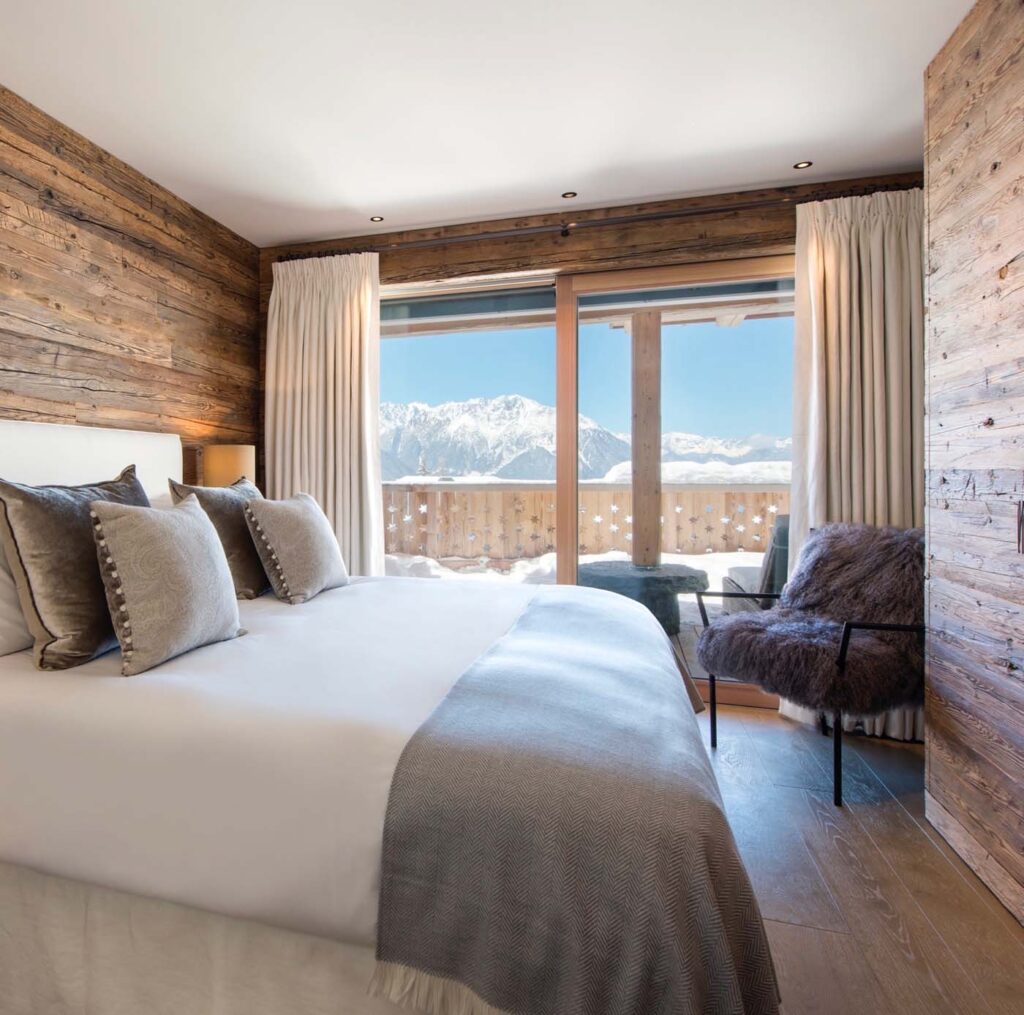 A cozy bedroom with wood-paneled walls, a large bed with gray bedding and pillows, a fur chair, and floor-to-ceiling windows showing a snowy mountain view and a wooden balcony. David Pearson Travel