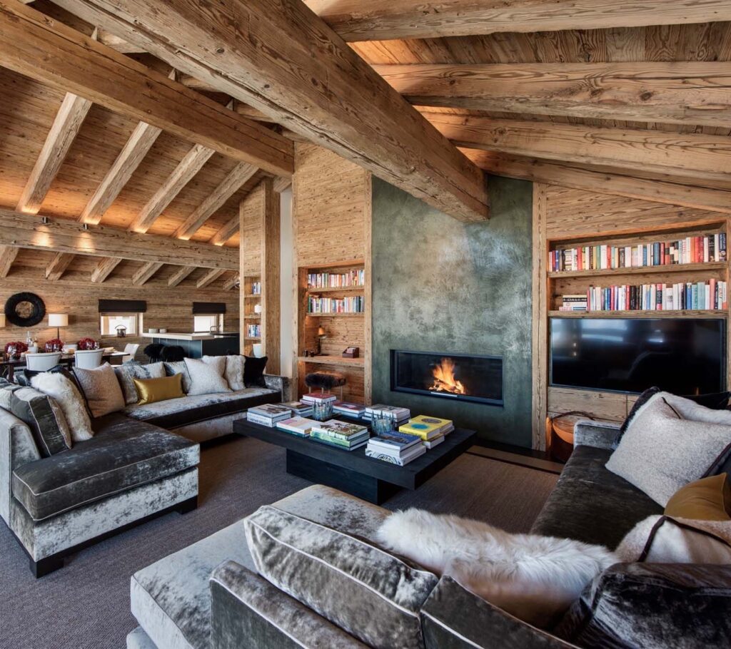 A cozy living room with exposed wooden beams, built-in bookshelves, a modern fireplace, plush velvet sofas, and a large coffee table. Warm lighting and neutral tones create a welcoming atmosphere. David Pearson Travel