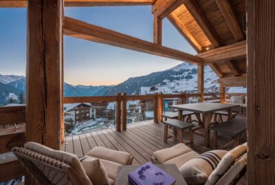 Cozy wooden balcony with lounge chairs, a table, and benches overlooking a snowy mountain village at sunset, with warm indoor lighting and scenic alpine views in the background. David Pearson Travel