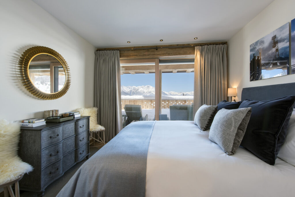 A cozy bedroom with a neatly made bed, gray and white bedding, a dresser with a round mirror, and large windows with mountain views. Two chairs sit on a balcony outside, and soft natural light fills the room. David Pearson Travel