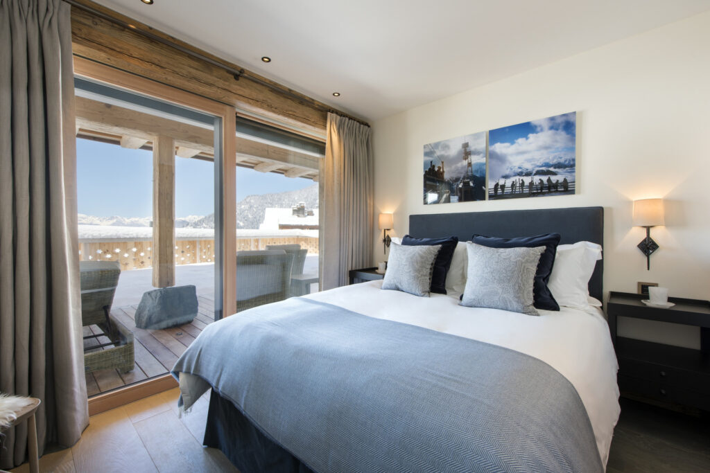 A cozy bedroom with a double bed, blue and white bedding, and large glass doors opening to a balcony with outdoor seating and mountain views. Neutral tones and soft lighting create a warm, inviting atmosphere. David Pearson Travel