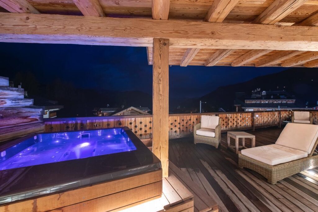 A rooftop terrace at night features a glowing hot tub with purple lights, wicker lounge chairs with cushions, and wooden beams and railings, overlooking mountains and buildings under a dark sky. David Pearson Travel