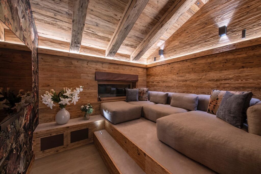 A cozy, wood-paneled room with exposed beams, tiered beige sectional seating, decorative pillows, soft lighting, and a flower vase on a low table, creating a warm, inviting atmosphere. David Pearson Travel