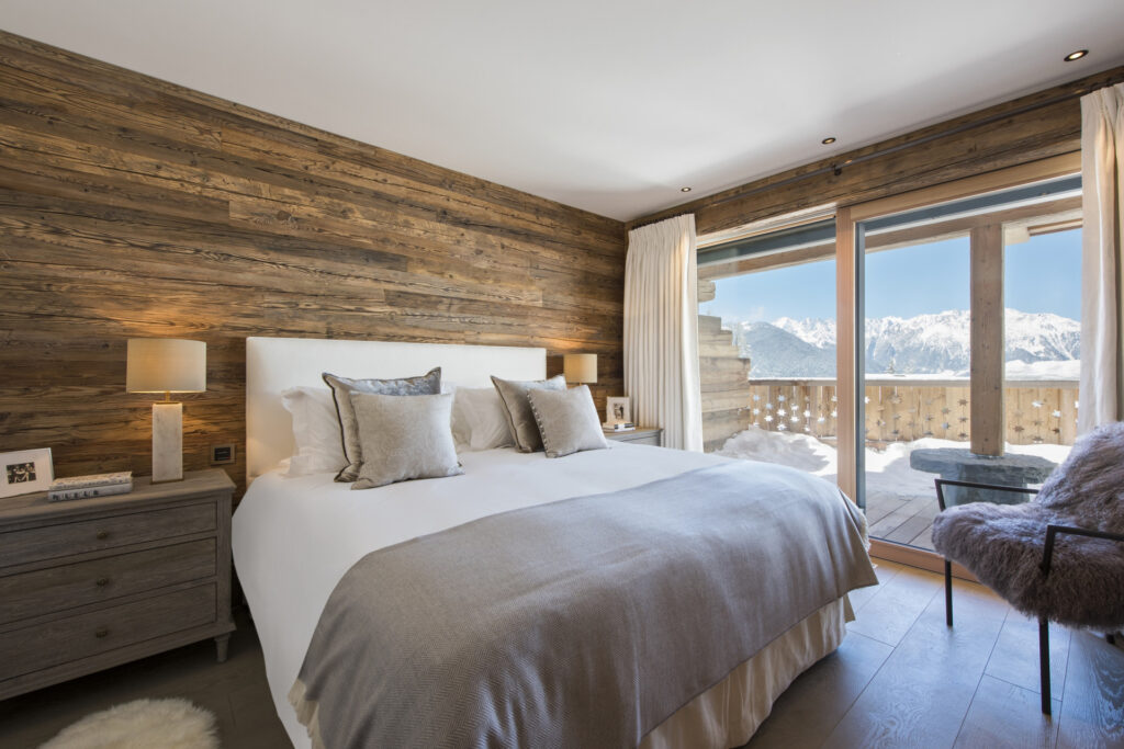 Modern bedroom with wood-paneled walls, a large bed with neutral bedding, two bedside tables with lamps, a cozy chair, and a balcony overlooking snowy mountains and a clear blue sky. David Pearson Travel
