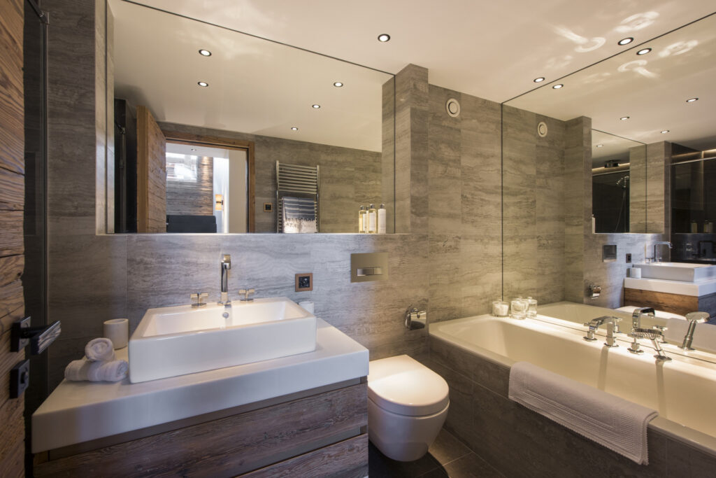 Modern bathroom with gray stone walls, a large mirror, rectangular sink, white countertop, toilet, and a bathtub with a towel draped over the edge. Recessed ceiling lights provide bright, even lighting. David Pearson Travel