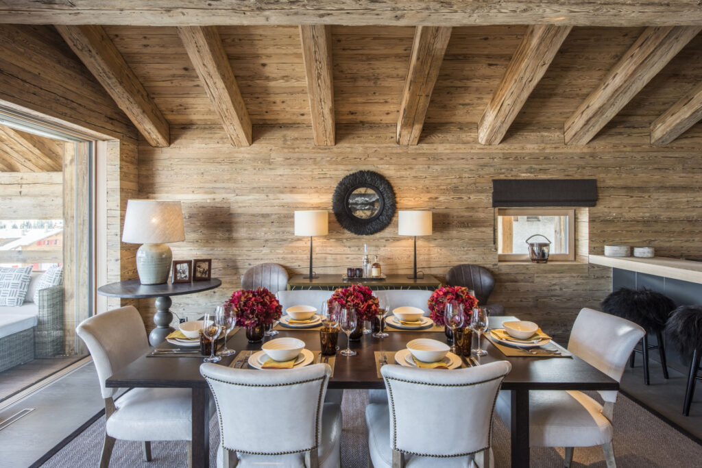 A rustic dining room with wood-paneled walls and exposed beams features a table set for six with white chairs, floral centerpieces, and elegant tableware. Soft lighting and decor elements create a cozy atmosphere. David Pearson Travel
