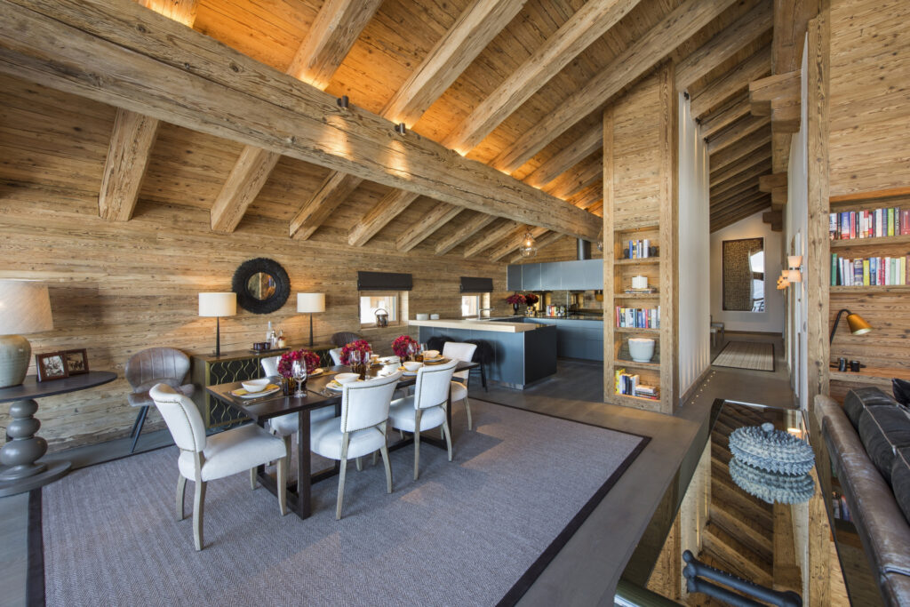 A spacious, modern cabin interior with exposed wooden beams, a dining table set for six, an open kitchen with sleek cabinets, bookshelves, and warm ambient lighting. The decor blends rustic and contemporary styles. David Pearson Travel