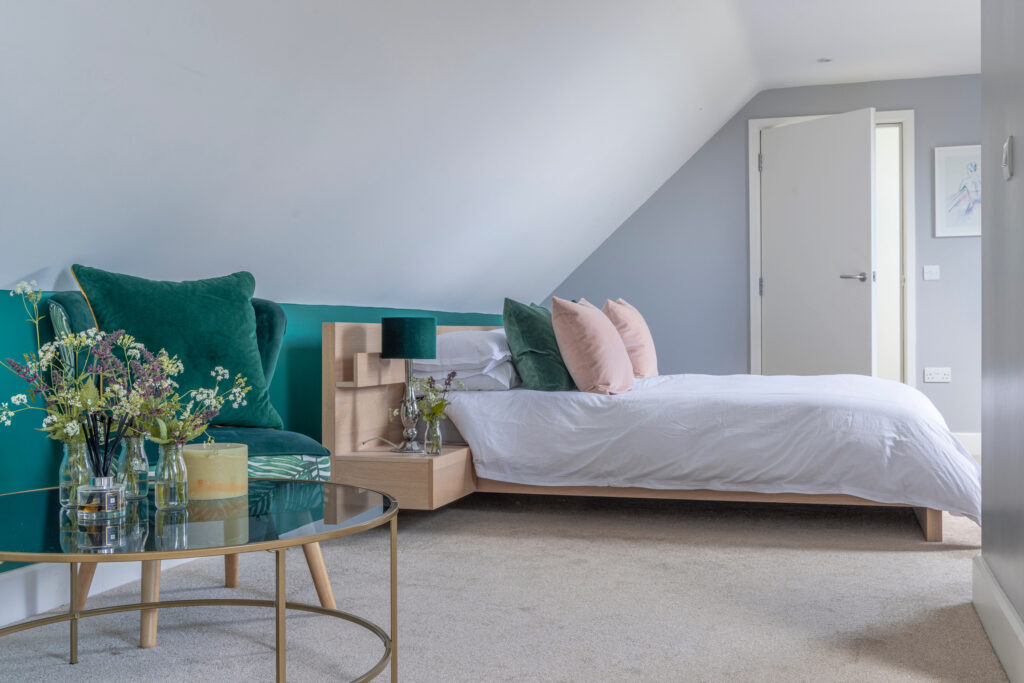 Modern bedroom with a sloped ceiling, neatly made bed with pink and green pillows, open door, glass coffee table with candles and flowers, and neutral carpet flooring. Walls are painted light gray and white. David Pearson Travel