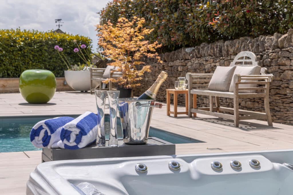 A spa area with a hot tub in the foreground, a bottle of champagne in an ice bucket with glasses, rolled blue and white towels, and a swimming pool beside a stone wall and garden seating under sunny skies. David Pearson Travel