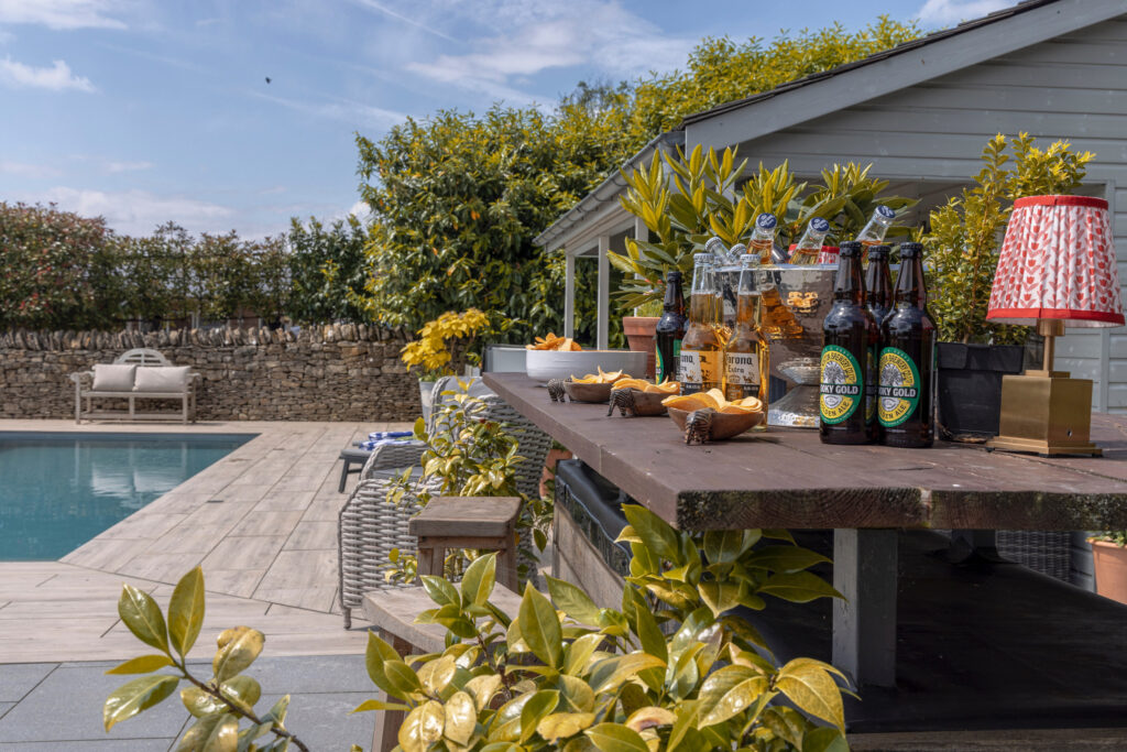 A poolside bar with bottles of beer, snacks, and drinks on a wooden counter, set in a sunny outdoor area with a swimming pool, greenery, and a stone wall in the background. David Pearson Travel