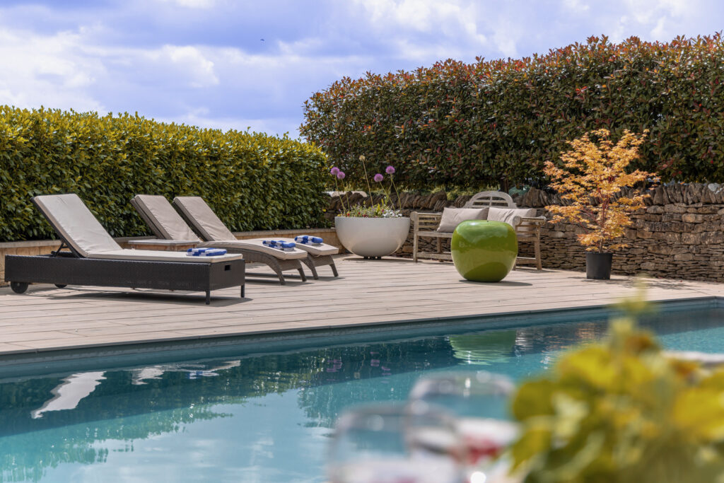 A sunny outdoor poolside area with lounge chairs, potted plants, a large green ceramic ornament, and a wooden bench, surrounded by tall hedges and a stone wall under a blue sky with clouds. David Pearson Travel