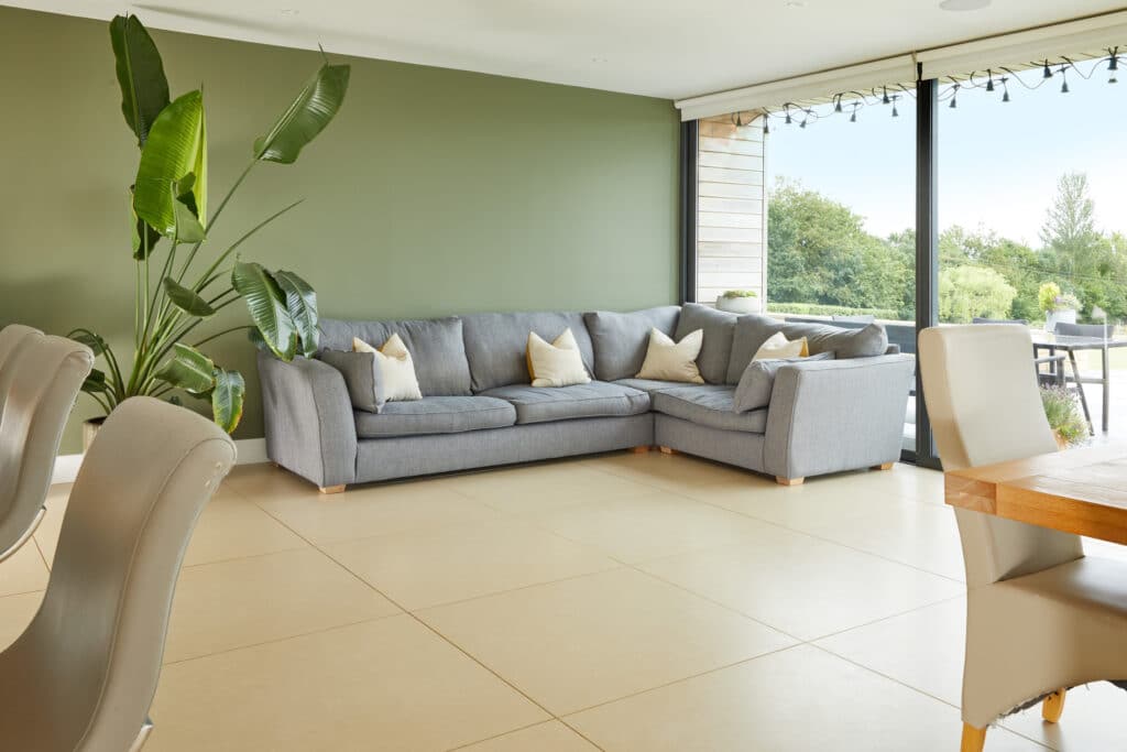 A modern living room with a large gray sectional sofa, cream pillows, a tall green plant, beige tile flooring, and floor-to-ceiling windows overlooking a garden. Dining chairs and a wooden table are partially visible. David Pearson Travel