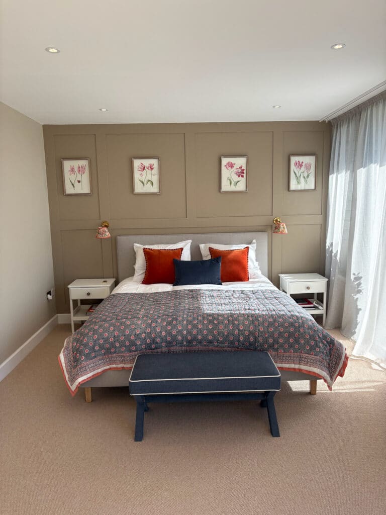 A neatly made bed with orange and navy accent pillows and a patterned quilt stands against a taupe paneled wall, flanked by white nightstands and floral artwork. Sheer curtains allow sunlight to brighten the bedroom. David Pearson Travel