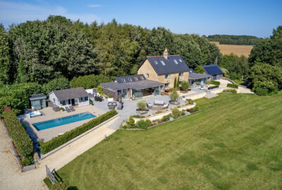Aerial view of a large countryside house with a stone exterior, surrounded by trees and fields. The property features a swimming pool, patio seating areas, outbuildings, landscaped gardens, and an expansive lawn. David Pearson Travel