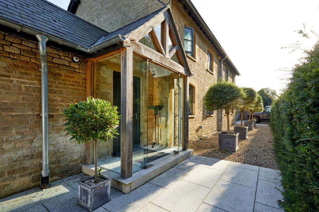 A modern glass entryway with wooden beams extends from a stone house. Potted topiary trees flank the entrance, and a gravel path runs alongside neatly trimmed hedges. The scene is bright and sunlit. David Pearson Travel