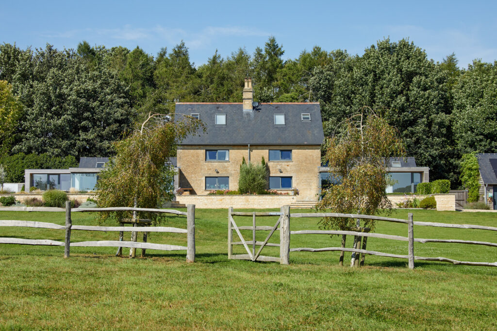 A stone house with a dark slate roof sits behind a rustic wooden fence on a green lawn, surrounded by trees under a clear blue sky. David Pearson Travel