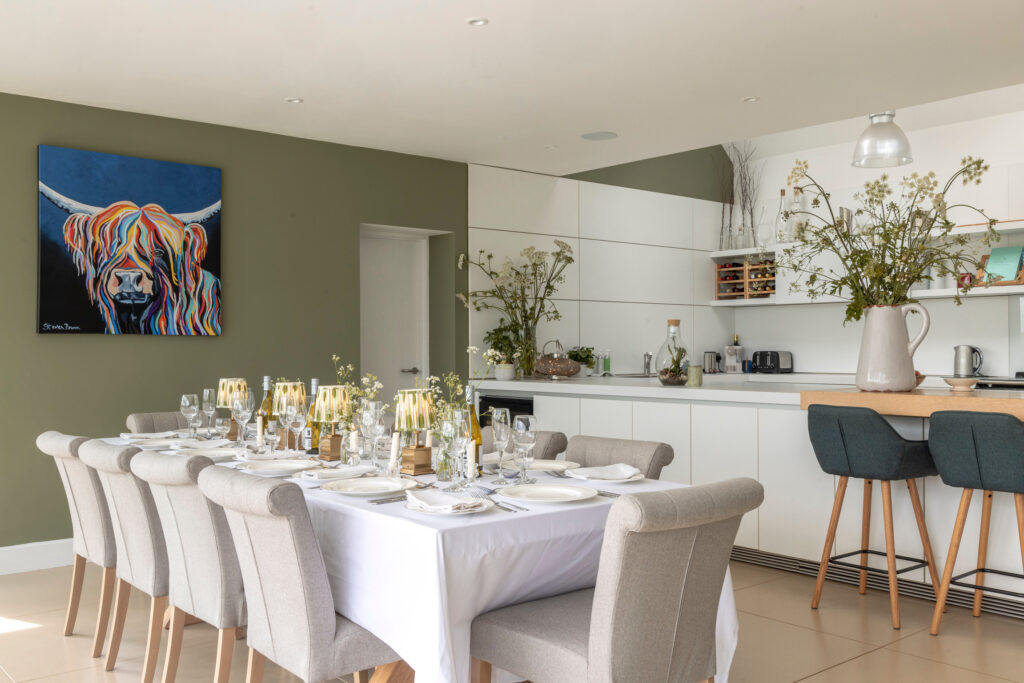 Modern dining room and kitchen with a set dining table for eight, neutral-toned chairs, white cabinets, green accent wall with colorful cow artwork, and floral arrangements on the table and counter. David Pearson Travel