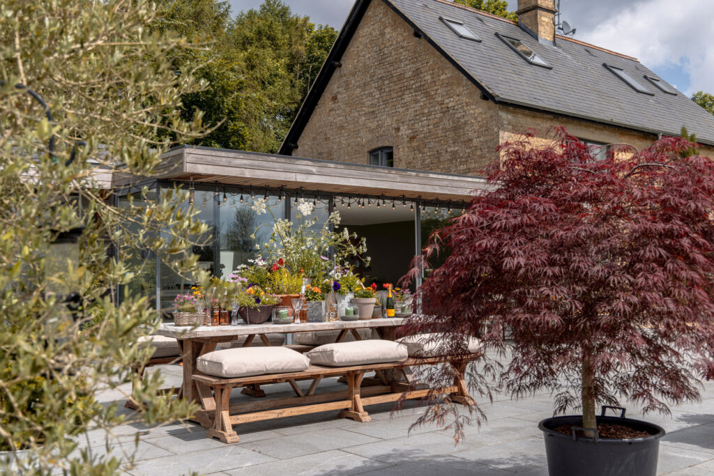 A rustic outdoor dining area with wooden benches and a table decorated with flowers sits on a stone patio beside a modern house, surrounded by greenery and a potted red-leafed tree. David Pearson Travel