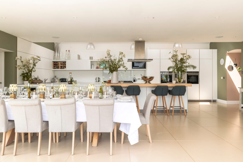 Modern open-plan kitchen and dining area with a long table set for a meal, beige chairs, a white tablecloth, and decorative plants; bar stools line a kitchen island in front of white cabinetry and built-in appliances. David Pearson Travel