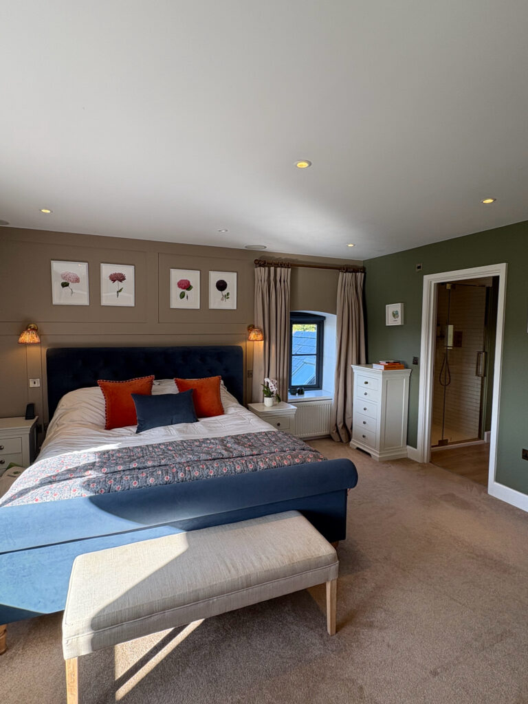 A cozy bedroom with a blue upholstered bed, white bedding, and orange accent pillows. Wall art with floral prints hangs above the bed. Theres a white bench, dresser, and an open door to a bathroom. A window with beige curtains lets in natural light. David Pearson Travel