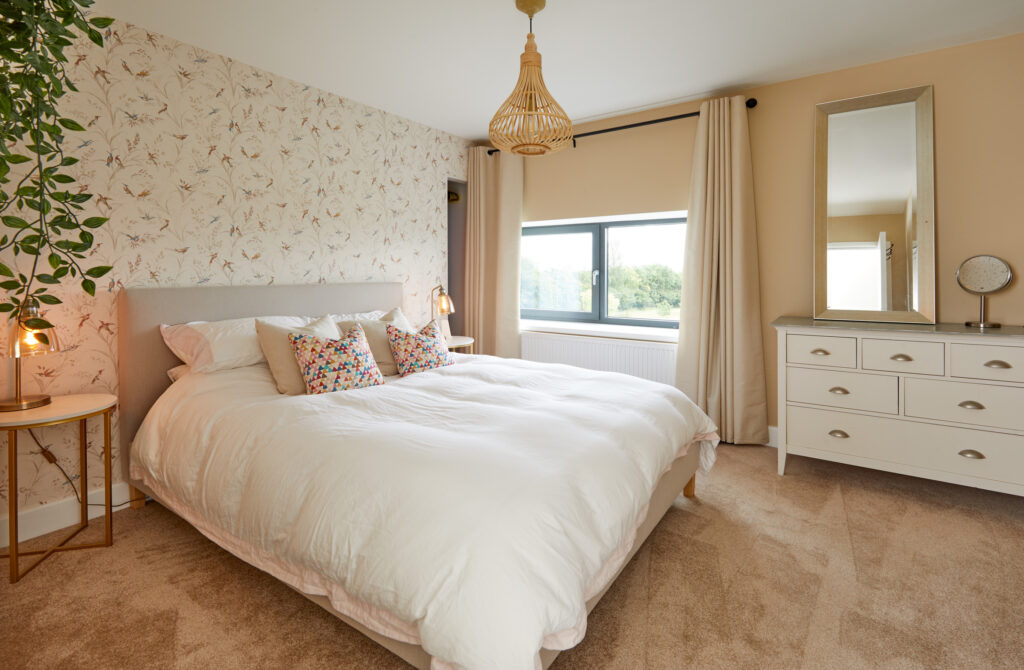 A cozy bedroom with a large bed, white bedding, colorful pillows, floral accent wall, beige carpet, dresser with a mirror, window with cream curtains, and hanging pendant light. A plant hangs in the corner. David Pearson Travel