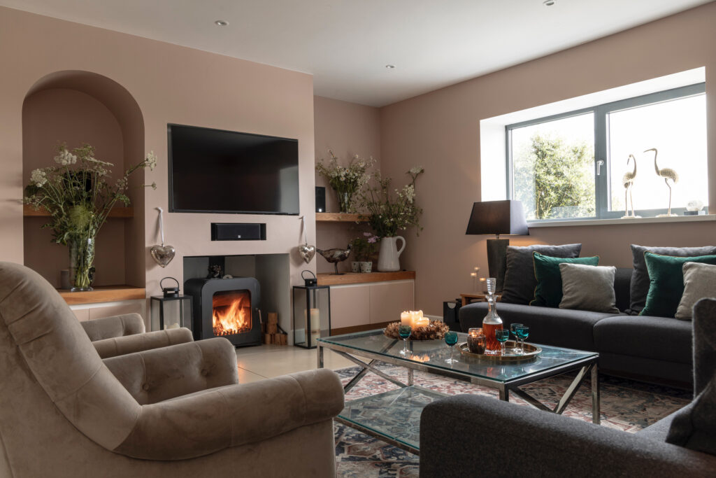 A cozy living room with neutral walls, a fireplace below a wall-mounted TV, gray sofas with green and gray cushions, candles on a glass coffee table, and natural light from a large window. David Pearson Travel