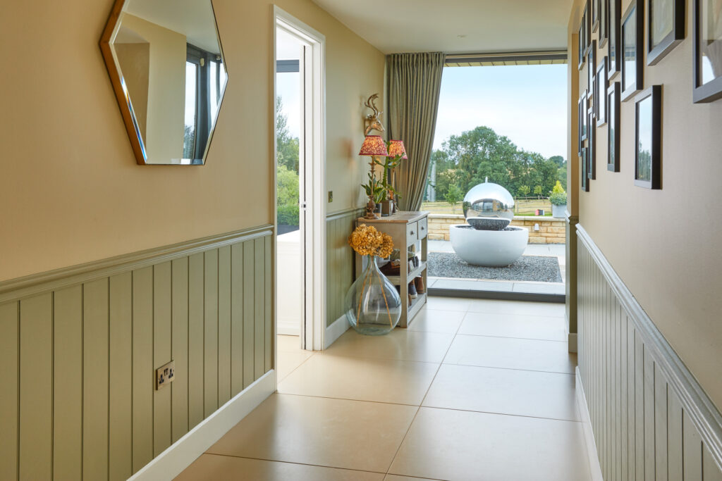 A bright hallway with light tiled floors, green panel walls, a hexagonal mirror, framed photos, and a console table with flowers. Glass doors open to a patio with a modern water feature and lush green scenery outside. David Pearson Travel