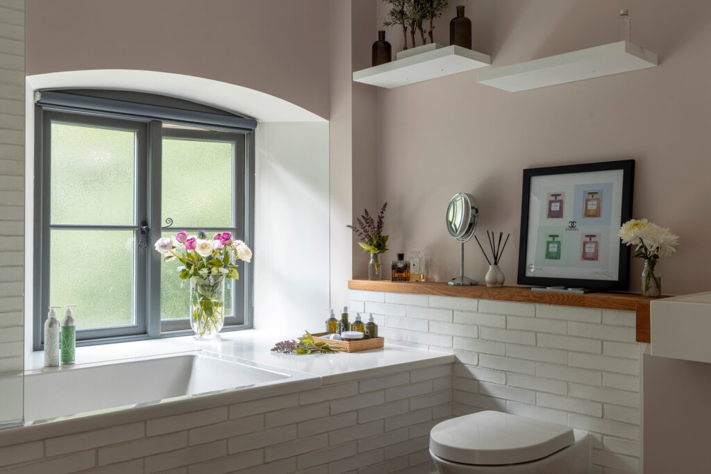 A modern bathroom with white subway tiles, a bathtub under a frosted window, fresh flowers in vases, decorative bottles, a small mirror, and framed art on a wooden shelf above the toilet. David Pearson Travel