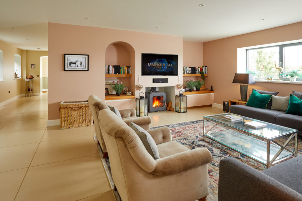 A modern living room with beige walls, a fireplace below a wall-mounted TV, bookshelves, two armchairs, a gray sofa, a glass coffee table, decorative plants, and a window letting in natural light. David Pearson Travel